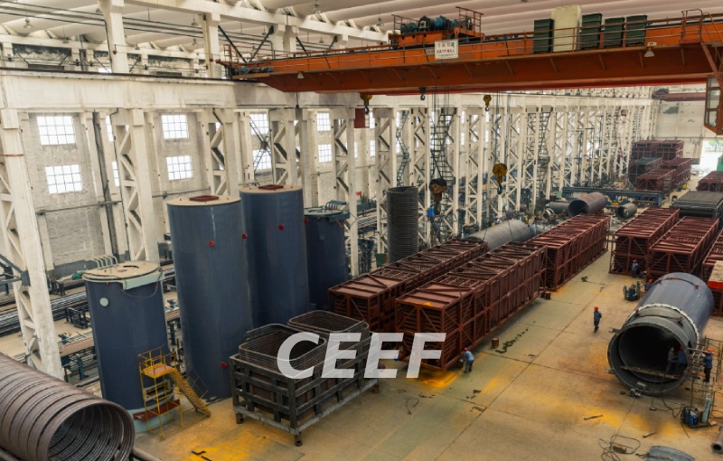 Interior of the CEEF heavy manufacturing workshop with an overhead bridge crane, vertical cylindrical thermal oil heater bodies and rows of modular convection tube banks under fabrication